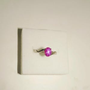 May include: A silver ring with a swirling band design, featuring a large, oval-shaped, pink gemstone with a star-like pattern. The ring is set against a white background.