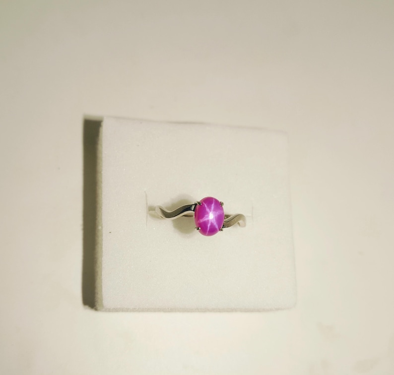 May include: A silver ring with a swirling band design, featuring a large, oval-shaped, pink gemstone with a star-like pattern. The ring is set against a white background.