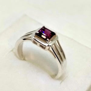 Könnte beinhalten: Ein silberner Ring mit einem rechteckigen, tiefvioletten Edelstein. Der Ring hat ein modernes Design mit einem Band, das sich auf jeder Seite des Edelsteinfassungs in drei parallele Linien aufteilt. Er liegt auf einer weißen Oberfläche.