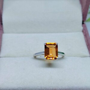 Könnte beinhalten: Ein silberner Ring mit einem großen, rechteckigen, bernsteinfarbenen Edelstein. Der Edelstein ist in einer einfachen Vier-Krappen-Fassung gefasst. Der Ring wird in einer rosa Schmuckschatulle präsentiert.