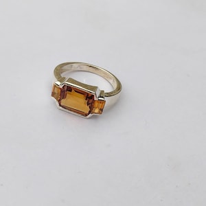 Könnte beinhalten: Ein silberner Ring mit einem rechteckigen, bernsteinfarbenen Edelstein als Mittelstück. Zwei kleinere, bernsteinfarbene Edelsteine flankieren den Hauptstein. Der Ring hat ein schlichtes, elegantes Design, das für den Alltag oder besondere Anlässe geeignet ist.