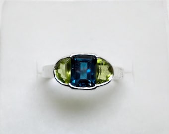 May include: A silver ring featuring a rectangular, deep blue gemstone flanked by two oval, light green gemstones. The ring's design is symmetrical, with the gemstones set in a polished silver band. A piece of jewelry.