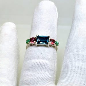 Könnte beinhalten: Ein silberner Ring mit einem rechteckigen, dunkelblauen Mittelstein, flankiert von kleineren roten und grünen Edelsteinen. Der Ring wird vor einem weißen Hintergrund präsentiert, um sein Design und seine Farben hervorzuheben.