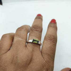 May include: A silver ring featuring a rectangular, light green gemstone. The ring is displayed on a finger against a white backdrop. The gemstone is a vibrant green, and the ring has a simple, modern design.