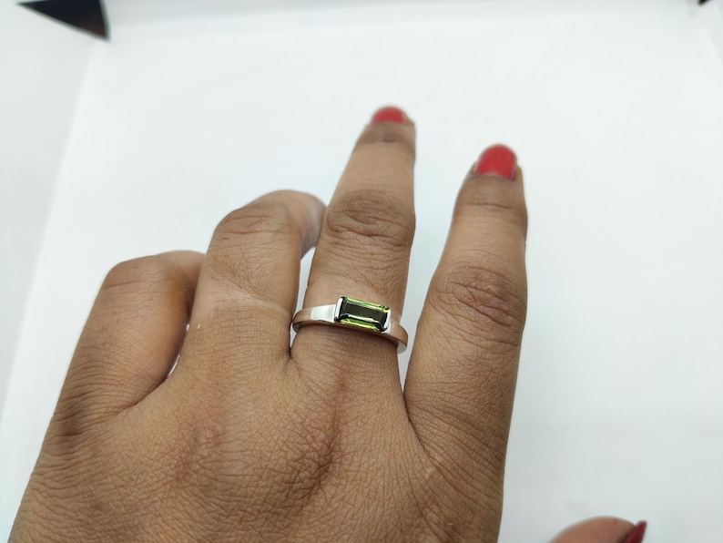 May include: A silver ring featuring a rectangular, light green gemstone. The ring is displayed on a finger against a white backdrop. The gemstone is a vibrant green, and the ring has a simple, modern design.