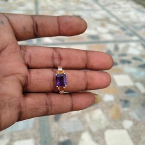 May include: A gold-toned ring featuring a rectangular, deep purple gemstone. The ring has a simple band with small, yellow-toned accents on either side of the gemstone. The ring is held in a hand, showcasing the jewelry.