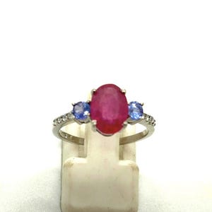 Könnte beinhalten: Ein silberner Ring mit einem großen ovalen Rubin-Mittelstein, flankiert von zwei kleineren blauen Edelsteinen. Das Ringband ist mit kleinen, klaren Akzentsteinen verziert, wodurch ein Dreistein-Design entsteht.