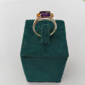 May include: A silver ring featuring a rectangular, deep purple gemstone as the centerpiece, flanked by two smaller, orange-yellow stones. The ring is displayed on a dark green velvet jewelry box, showcasing the ring's design and color contrast.
