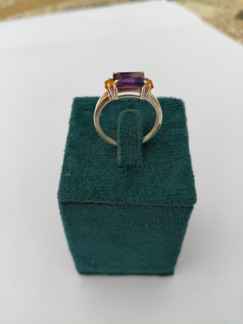 May include: A silver ring featuring a rectangular, deep purple gemstone as the centerpiece, flanked by two smaller, orange-yellow stones. The ring is displayed on a dark green velvet jewelry box, showcasing the ring's design and color contrast.