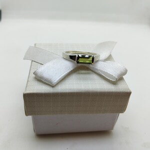 May include: A silver ring with a rectangular, light green gemstone sits atop a white gift box, adorned with a white ribbon. The ring's design is simple yet elegant. The box is a neutral colour, providing a clean backdrop for the jewellery.