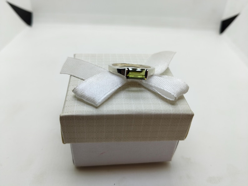 May include: A silver ring with a rectangular, light green gemstone sits atop a white gift box, adorned with a white ribbon. The ring's design is simple yet elegant. The box is a neutral colour, providing a clean backdrop for the jewellery.