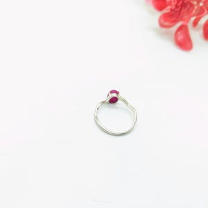 May include: A silver ring with a small, oval-shaped, red gemstone. The ring has a simple, elegant design with a slightly textured band. The background is white, with a few red flower petals in the upper right corner.