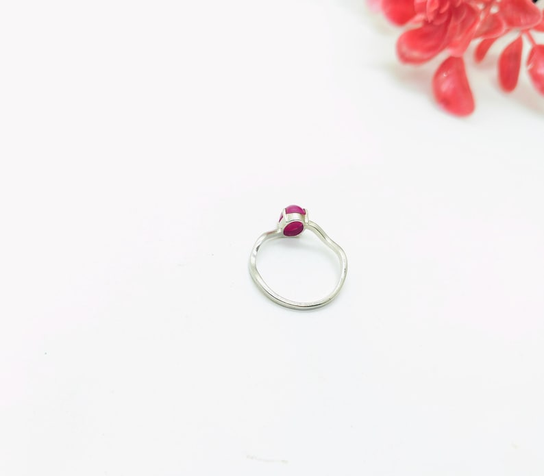 May include: A silver ring with a small, oval-shaped, red gemstone. The ring has a simple, elegant design with a slightly textured band. The background is white, with a few red flower petals in the upper right corner.
