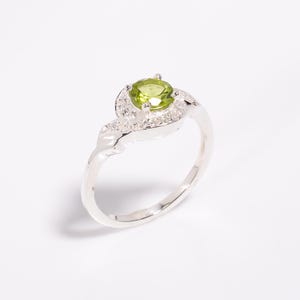 Könnte beinhalten: Silberring mit einem runden, hellgrünen Edelstein in der Mitte, umgeben von kleineren, klaren Steinen. Das Band des Rings hat ein wirbelndes Design, wodurch ein elegantes Schmuckstück entsteht.