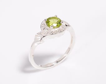 May include: A silver ring with a central, round, light green gemstone, encircled by smaller, clear stones. The ring's band has a swirling design. A stylish piece of jewellery.