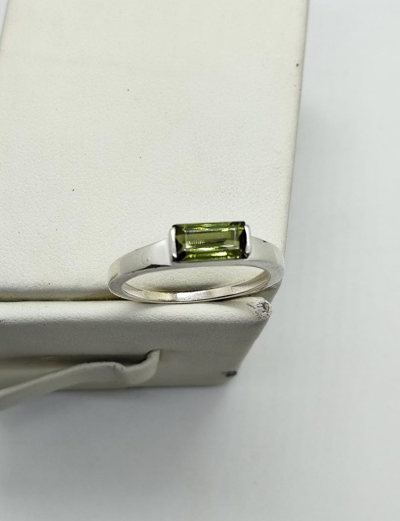 May include: A silver ring with a rectangular, light green gemstone. The gemstone is set in a simple, modern band. The ring is displayed on a white surface, highlighting its polished finish and elegant design.