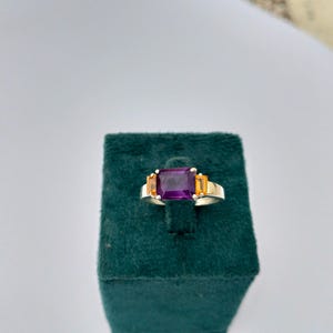 May include: A silver ring with a large rectangular purple gemstone as the centerpiece, flanked by two smaller orange gemstones. The ring is set on a dark green velvet surface, showcasing the jewelry's design and color contrast.