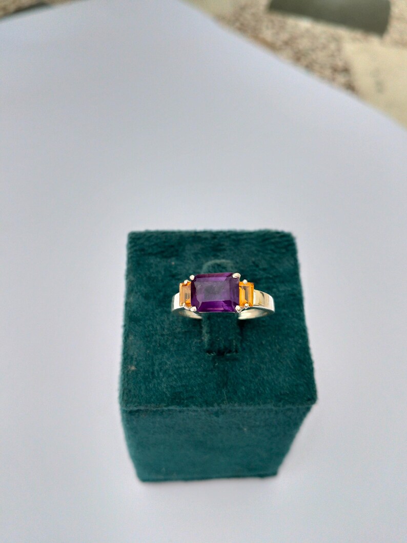 May include: A silver ring with a large rectangular purple gemstone as the centerpiece, flanked by two smaller orange gemstones. The ring is set on a dark green velvet surface, showcasing the jewelry's design and color contrast.