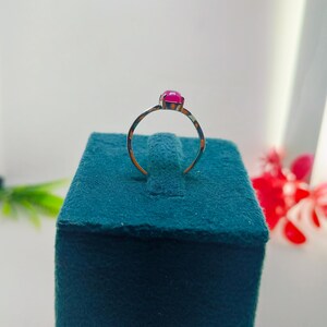 May include: A gold-colored ring with a vibrant, round, magenta gemstone. The ring is set on a teal velvet surface, with a white background and hints of red flowers. The ring's band is thin and delicate.