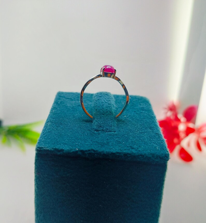 May include: A gold-colored ring with a vibrant, round, magenta gemstone. The ring is set on a teal velvet surface, with a white background and hints of red flowers. The ring's band is thin and delicate.