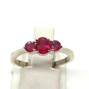 Könnte beinhalten: Ein silberfarbener Ring mit einem zentralen, oval geschliffenen, leuchtend roten Edelstein, der von zwei kleineren, rund geschliffenen roten Edelsteinen flankiert wird. Die Ringschiene ist glatt und metallisch und hält die drei Steine in einer Krappenfassung.