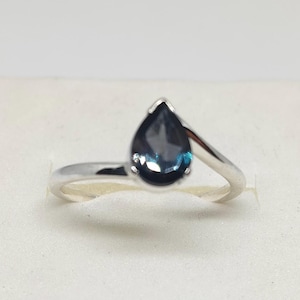 Könnte beinhalten: Ein silberner Ring mit einem tropfenförmigen, dunkelblauen Edelstein. Der Edelstein hat einen leichten Türkis-Farbton und ist in einer Zargenfassung gefasst. Das Band umschließt den Stein und erzeugt ein modernes, elegantes Design.