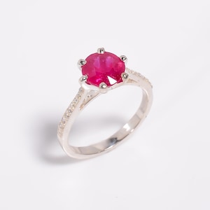 Könnte beinhalten: Ein silberner Ring mit einem großen, runden, leuchtend roten Edelstein im Mittelpunkt. Das Band ist mit kleineren, klaren Edelsteinen besetzt. Der Ring ist vor einem schlichten weißen Hintergrund platziert.