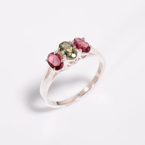 Könnte beinhalten: Ein silberner Ring mit drei ovalen Edelsteinen in Rosa- und Grüntönen. Die Edelsteine sind in einer Reihe über der Oberseite des Rings angeordnet. Der Ring ist vor einem weißen Hintergrund abgebildet.