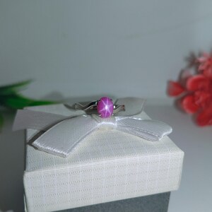 May include: A silver ring featuring a large, oval-cut, pink star sapphire gemstone. The ring is set on a white gift box with a white ribbon. The background includes red and green decorative elements.
