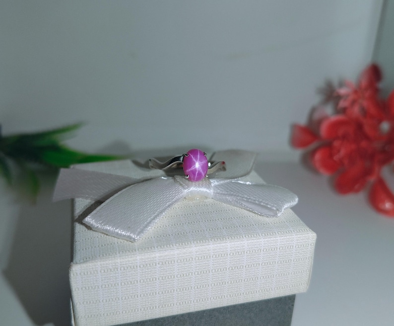 May include: A silver ring featuring a large, oval-cut, pink star sapphire gemstone. The ring is set on a white gift box with a white ribbon. The background includes red and green decorative elements.