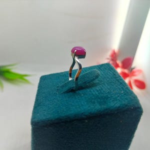 May include: A silver ring with a large, oval-shaped, magenta gemstone. The ring's band has a unique, curved design. The ring is displayed on a teal-colored, textured surface, with a blurred background.