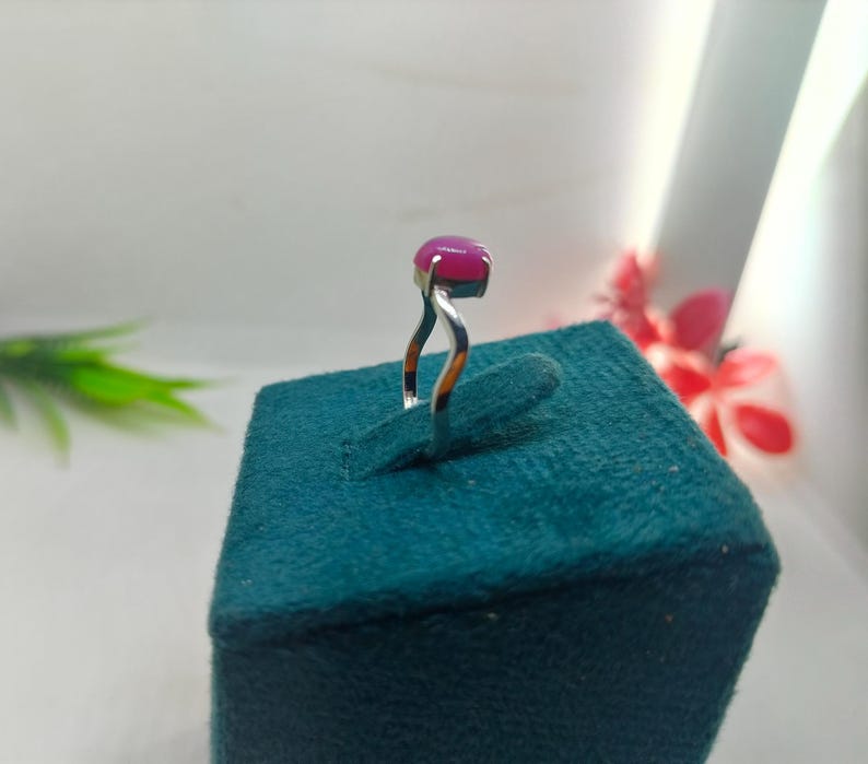 May include: A silver ring with a large, oval-shaped, magenta gemstone. The ring's band has a unique, curved design. The ring is displayed on a teal-colored, textured surface, with a blurred background.