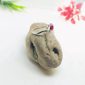 May include: A silver ring with a vibrant, square-cut, magenta gemstone. The ring is displayed on a textured, light brown stone against a white background. The ring's band has a simple, elegant design.