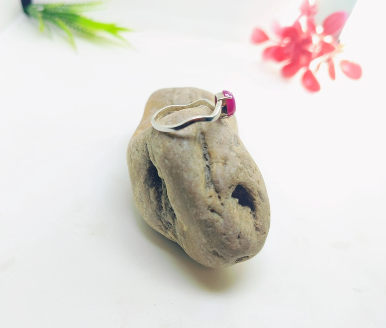 May include: A silver ring with a vibrant, square-cut, magenta gemstone. The ring is displayed on a textured, light brown stone against a white background. The ring's band has a simple, elegant design.
