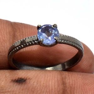 Könnte beinhalten: Ein Ring mit einem runden, hellblauen facettierten Edelstein in einer Vier-Krappen-Fassung. Die Ringschiene besteht aus dunklem, oxidiertem Metall und weist ein texturiertes, perlenartiges Kantendesign auf, das beidseitig entlang der Schiene verläuft.