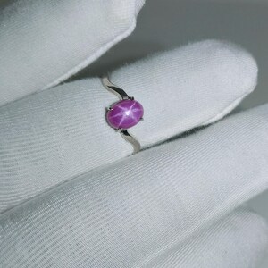 May include: A sterling silver ring featuring a vibrant, oval-shaped, star ruby gemstone. The ruby is a deep pink color with a six-ray star pattern. The ring's band has a unique, wavy design.