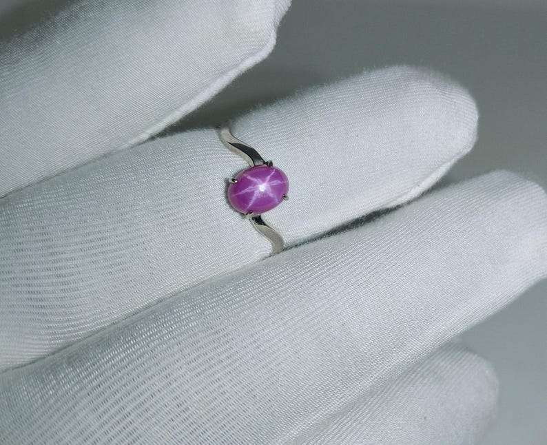 May include: A sterling silver ring featuring a vibrant, oval-shaped, star ruby gemstone. The ruby is a deep pink color with a six-ray star pattern. The ring's band has a unique, wavy design.