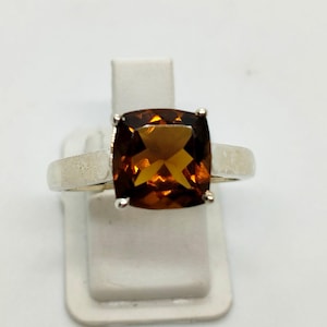Könnte beinhalten: Ein silberner Ring mit einem großen, quadratisch geschliffenen, bernsteinfarbenen Edelstein. Der Edelstein ist in einem schlichten, modernen Design gefasst, das seine warmen, goldenen Farbtöne hervorhebt. Der Ring wird auf einem weißen Ständer präsentiert.