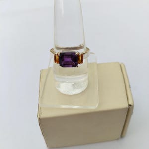 May include: A gold-toned ring featuring a large, rectangular, deep purple gemstone as the centerpiece. Two smaller, orange-toned gemstones flank the main stone. The ring is displayed on a clear, plastic ring holder.