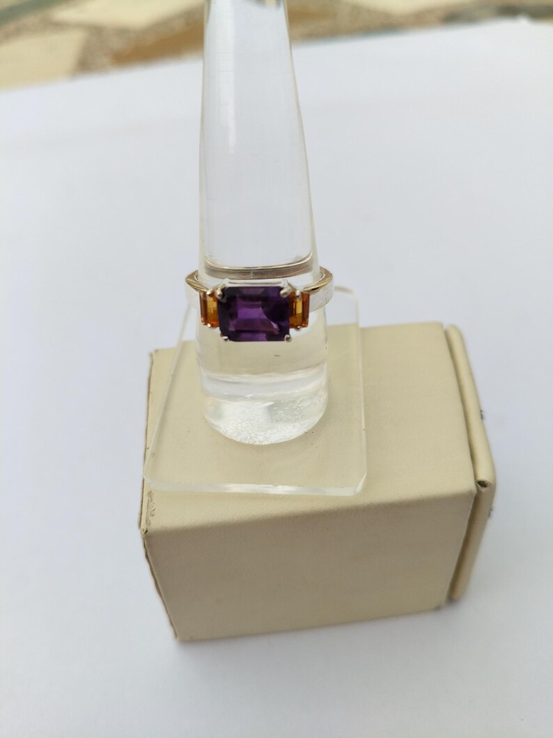 May include: A gold-toned ring featuring a large, rectangular, deep purple gemstone as the centerpiece. Two smaller, orange-toned gemstones flank the main stone. The ring is displayed on a clear, plastic ring holder.