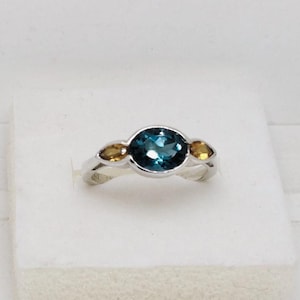 Könnte beinhalten: Ein silberner Ring mit einem großen ovalen, teal-farbenen Edelstein, flankiert von zwei kleineren, gelb-orangenen Edelsteinen. Das Band des Rings hat ein verdrehtes Design, und die Edelsteine sind in einer Zargenfassung gefasst. Dies ist ein Schmuckstück.