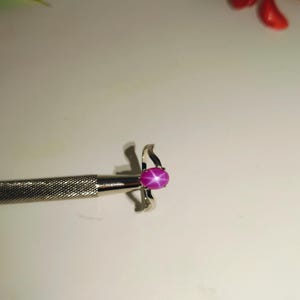 May include: A silver ring with a vibrant pink oval gemstone. The gemstone displays a star-like effect. The ring is held by a silver tool, showcasing the jewelry's design and craftsmanship. The ring is a piece of fine jewelry.