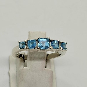 Könnte beinhalten: Ein silberfarbener Ring mit fünf quadratisch geschliffenen, hellblauen Edelsteinen in einer Reihe. Der mittlere Stein ist der größte, flankiert von zwei kleineren Steinen auf jeder Seite, gefasst in eine polierte Metallschiene.