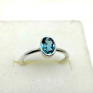 Könnte beinhalten: Ein silberner Ring mit einem hellblauen, ovalen Edelstein. Der Ring hat ein schlichtes Band und eine Fassung, die den Edelstein hält. Der Edelstein hat einen leuchtenden Blauton. Der Ring wird auf einer weißen Oberfläche präsentiert.