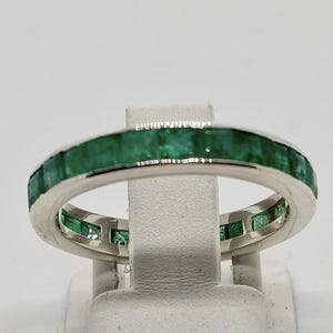 Könnte beinhalten: Ein silberfarbener Eternity-Ring mit einer durchgehenden Reihe quadratisch geschliffener, tiefgrüner Smaragd-Edelsteine, die in eine polierte Metallschiene eingefasst sind. Der Ring zeigt ein einheitliches Design mit Steinen rundherum.