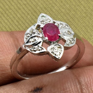 Könnte beinhalten: Ein silberner Ring mit einem ovalen Rubin in der Mitte, umgeben von einem floralen Design mit kleinen, funkelnden Diamanten. Der Ring hat ein glattes, poliertes Band, das die Edelsteine hervorhebt.