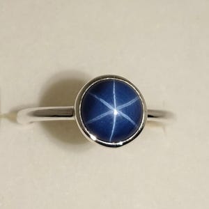 Könnte beinhalten: Ein silberner Ring mit einem runden, tiefblauen Edelstein, der ein sechszackiges Sternmuster aufweist. Der Edelstein ist in einer Lünettenfassung gefasst, und das Band ist aus poliertem Silber. Der Ring wird vor einem neutralen Hintergrund gezeigt.