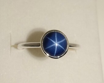 May include: A silver ring with a round, dark blue gemstone displaying a six-pointed star. The gemstone is set in a bezel, and the band is polished silver. The ring is shown against a plain background. The ring is a piece of jewellery.