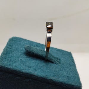 May include: A silver ring featuring a dark, rectangular gemstone. The ring is presented on a teal display. The band is slender and polished. The gemstone appears to be cut. The ring is angled to highlight its design.