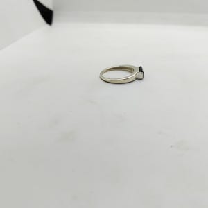May include: A silver ring featuring a rectangular black gemstone. The ring is displayed on a white surface. The band is slender and polished. The black stone is small and square.